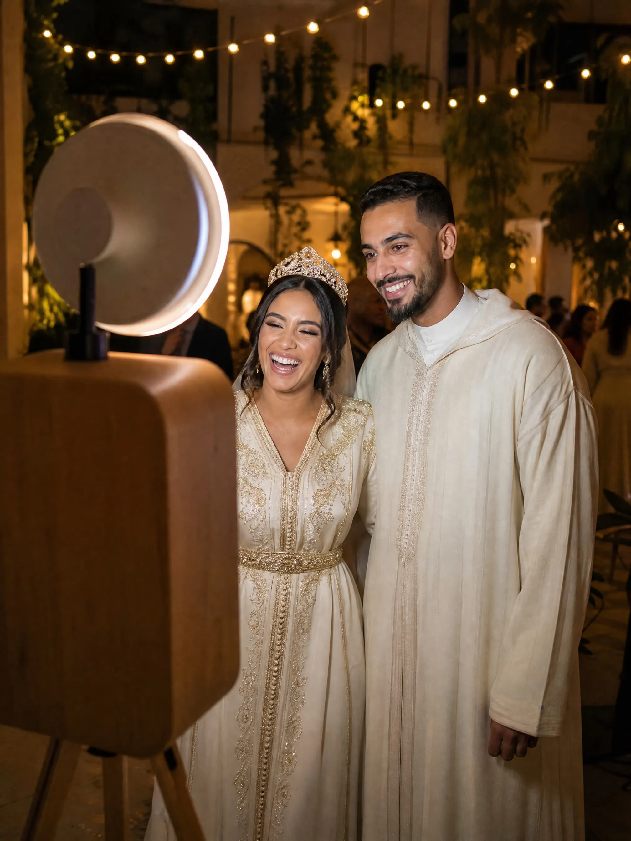 Invités en caftan qui rient devant le Photobooth Classique, borne en bois et couronne lumineuse allumée